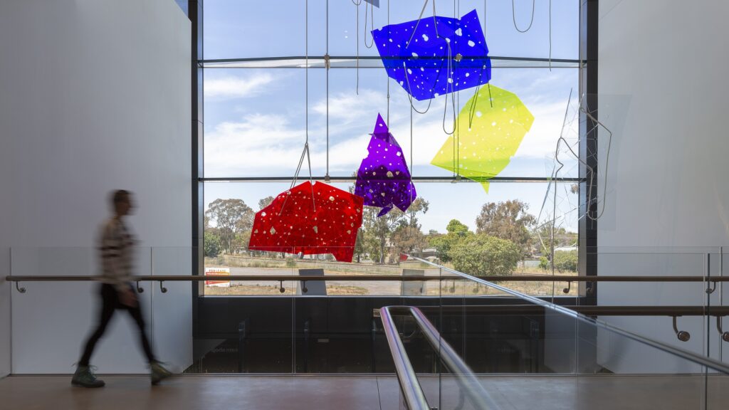 A photo of large colourful transluscent acrylic sculptures hang from the ceiling over a building's balustrade; a person is walking to their left.
