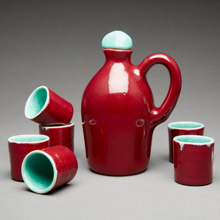 Photo of deep berry-red coloured ceramic cups and pitcher with aqua-blue interior glaze arranged against a grey backdrop.