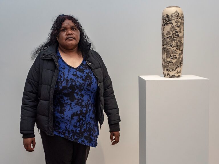 A woman in a blue shirt and black puffer jacket stands next to her award-winning ceramic vase