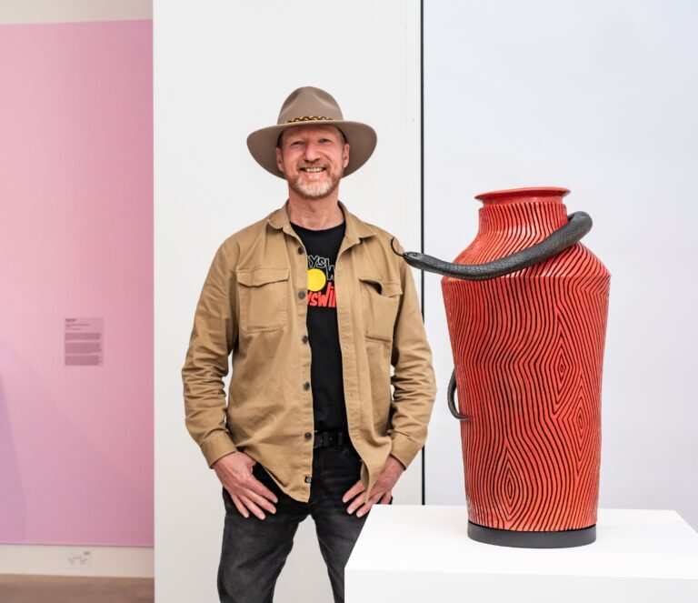 A man in an Akubra hat, tan jacket and black t-shirt decorated with the Aboriginal flag stands beside his award-winning ceramic vase
